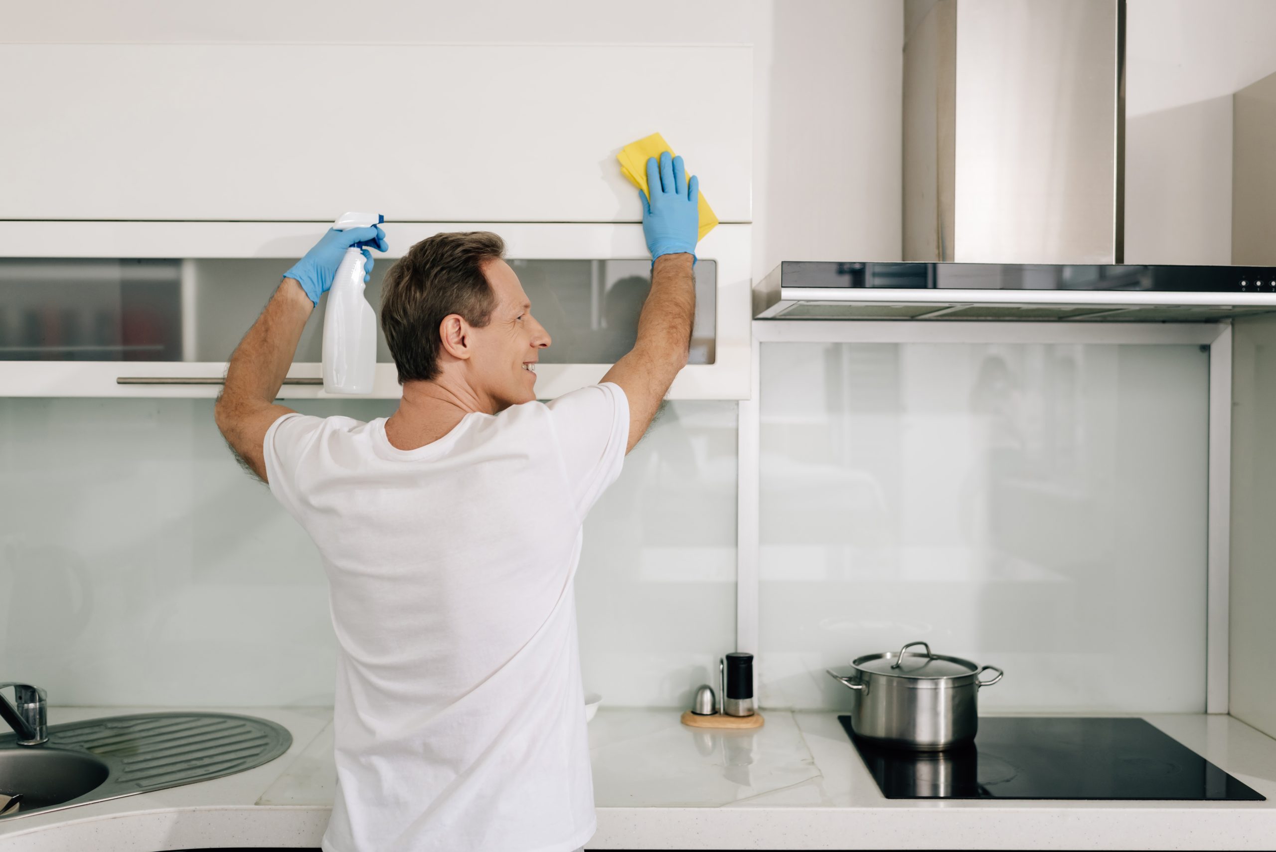 happy man in rubber gloves holding spray bottle and rag while cleaning kitchen