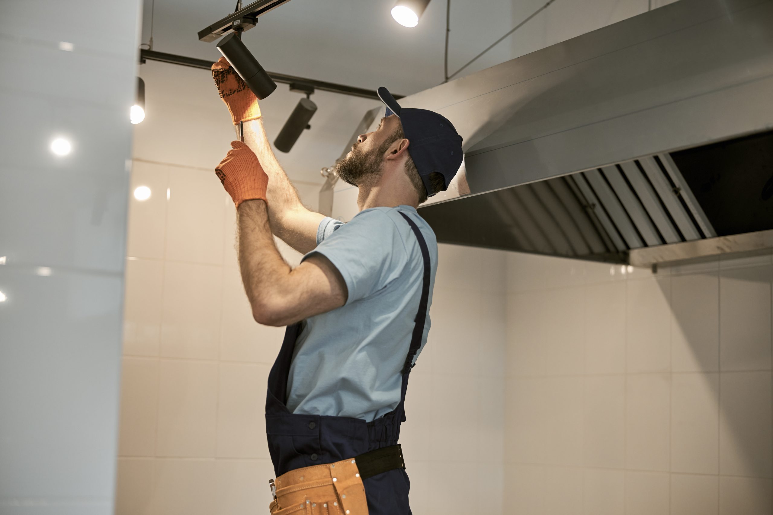 Male worker wearing work gloves and cap while repairing ceiling lamp with screwdriver