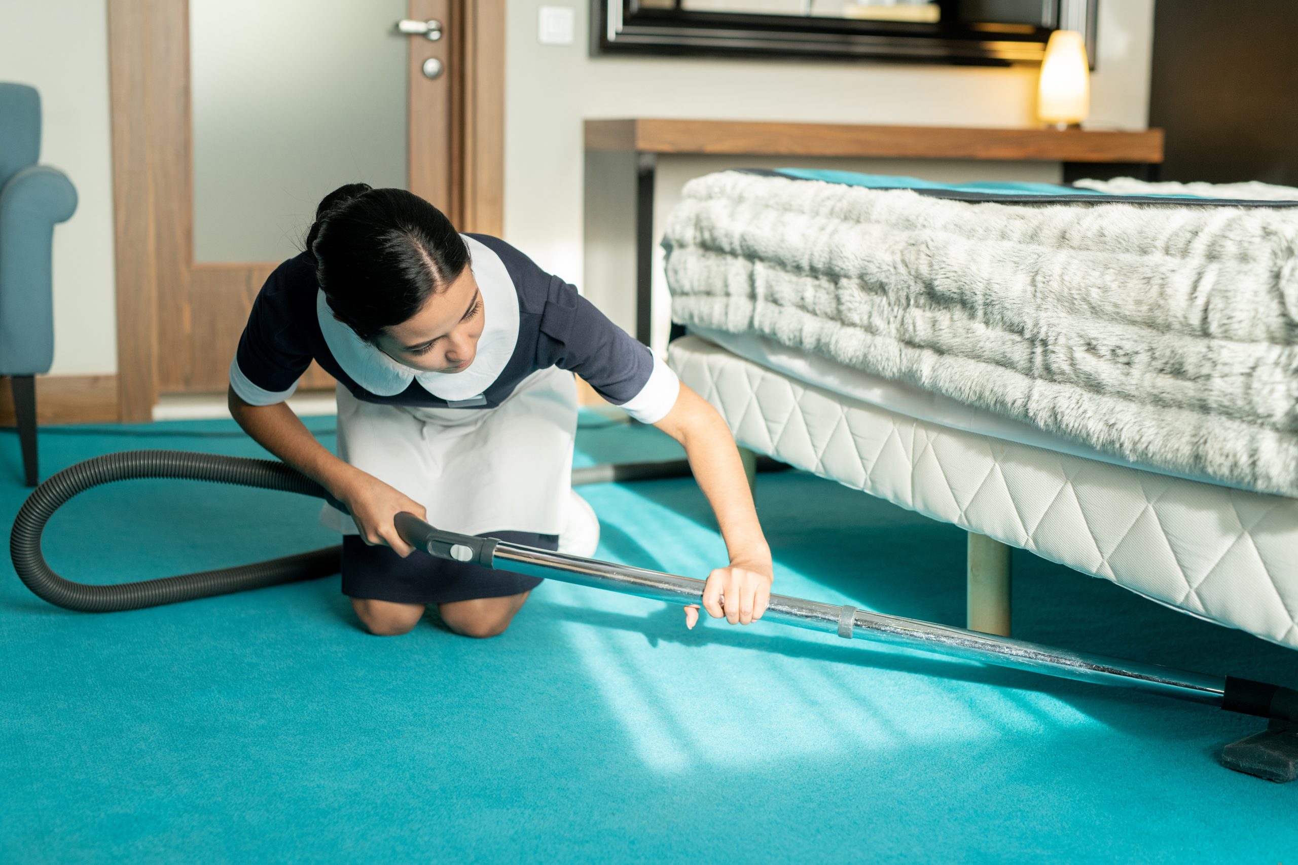 Young beautiful brunette chambermaid in uniform using vacuum cleaner while cleaning blue floor covering under bed in hotel room